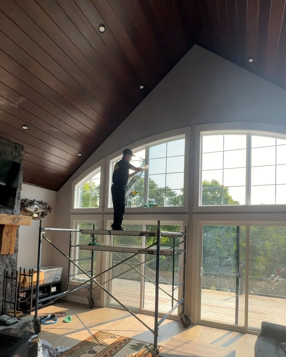 An installer on a rolling scaffold working on the arched upper windows of a vaulted cottage great room, with a wall of sliding doors opening to the deck below