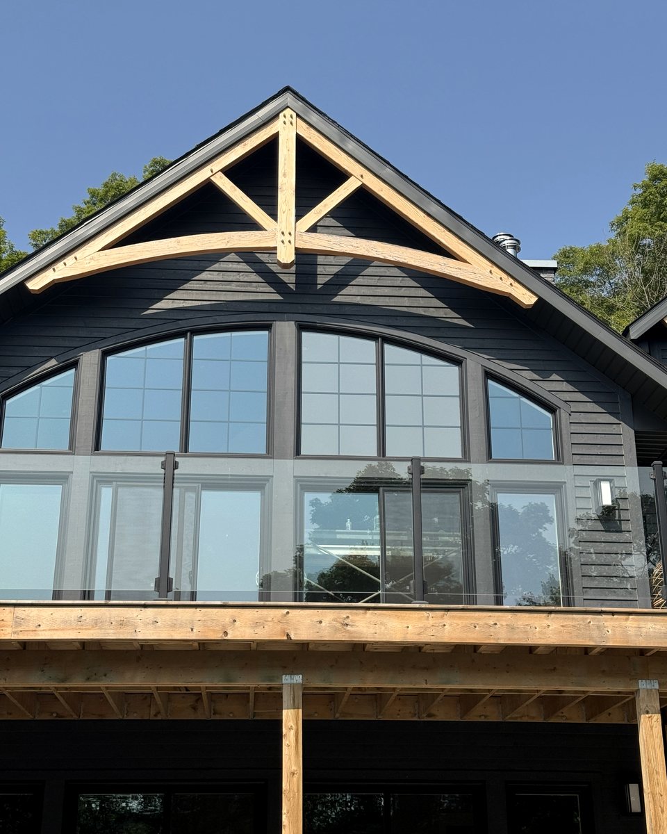 Timber-truss gable on a dark-sided cottage, with a row of arched upper windows above a glass-railed deck under a clear blue sky