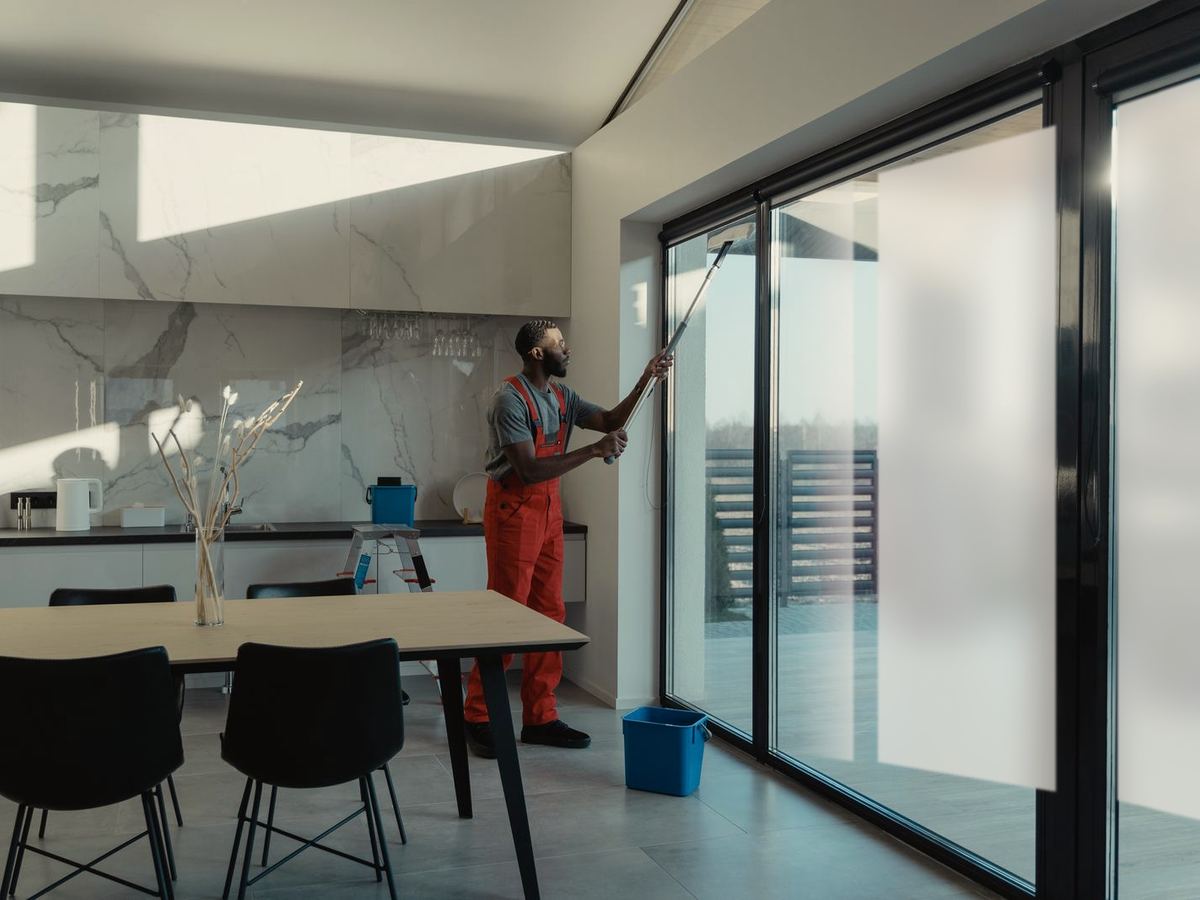 Technician working a floor-to-ceiling glass wall with his applicator as the switchable layer changes state, far panes frosted while the pane at his tool stays clear